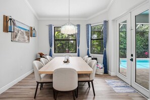 A dining room with a large table, chairs, a chandelier, and a view of a swimming pool through large windows.