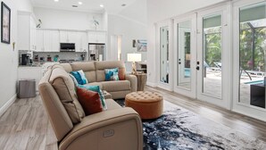 A spacious living room with a beige sofa, ottoman, and a view of a pool through glass doors. - Amazing Location - Walk to the Beach & JW ~Fully Loaded Inside & Out w/Pool, Beach Gear, WiFi & More (Marco Island)