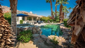 A pool area with a house in the background, surrounded by palm trees and rocks. - Gorgeous Home w/Saltwater Pool/Spa with MISTERS! PGA West - Fabulous Outdoor Setting-3BD#245594 (La Quinta)