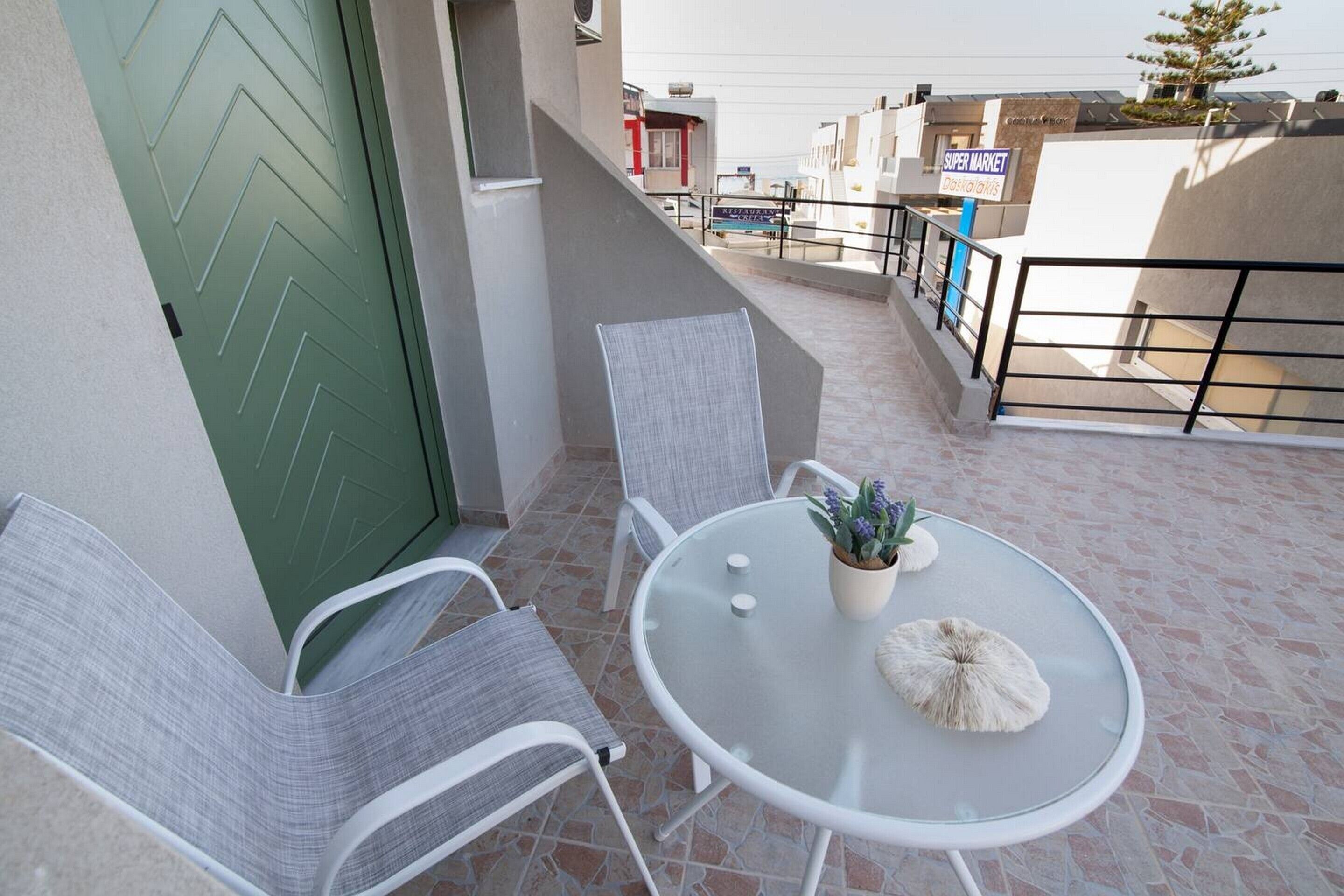 A balcony with a green door, two white chairs, a glass table, and a potted plant.