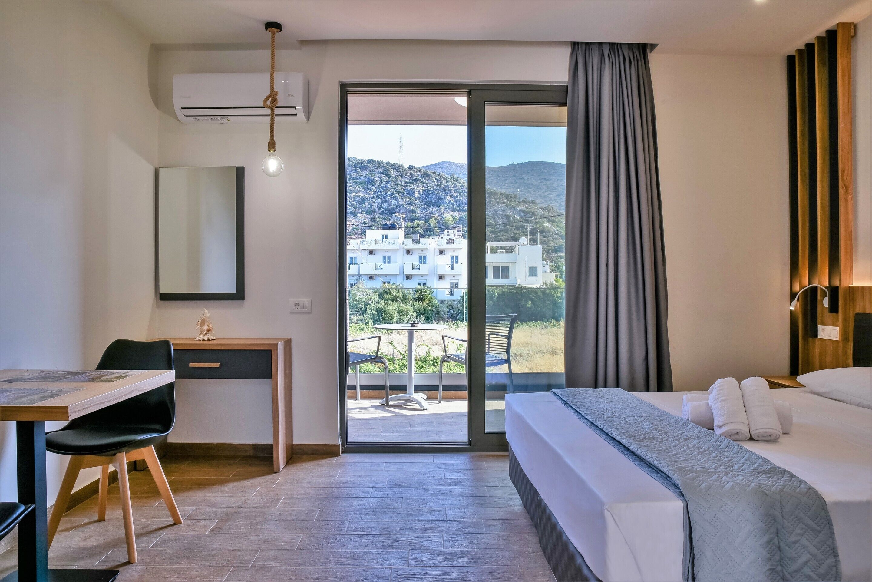Habitación de hotel con cama, escritorio, silla y vistas a edificios y colinas a través del ventanal.