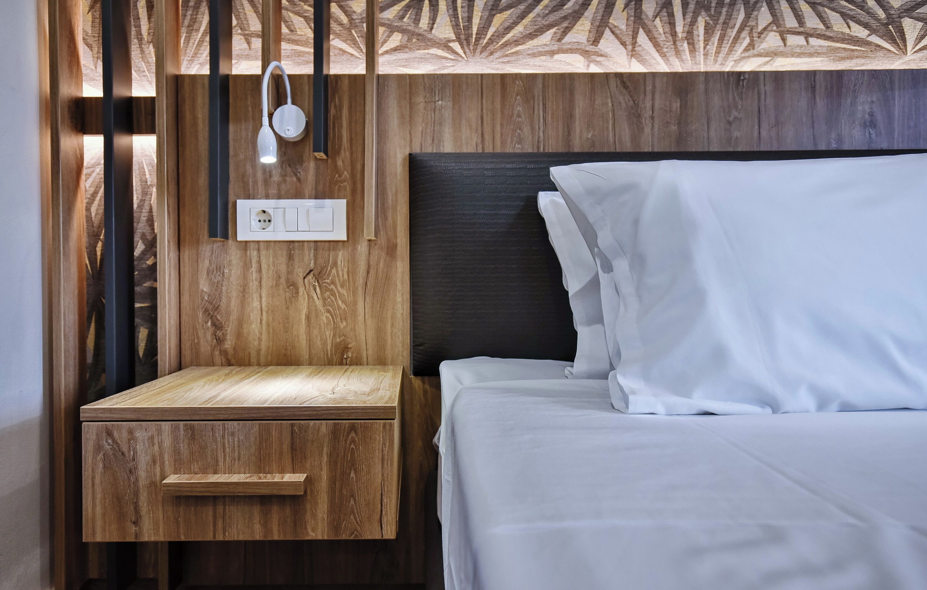 Una cama bien hecha con sábanas blancas, una mesita de noche de madera con un cajón y una lámpara fijada a la pared.