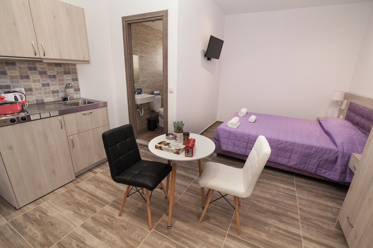 Une pièce de taille modeste comprenant un coin cuisine, un lit recouvert d’une courtepointe violette, deux chaises et une petite table sur laquelle sont posés divers objets.