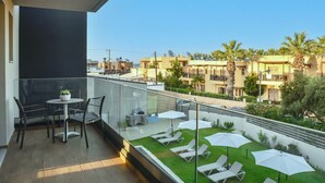 A balcony with a table and chairs, a small garden area with sun loungers and umbrellas, and a view of neighboring buildings. - Del Mar Apartments in Stalis (Hersonissos)