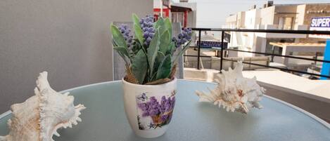 Una planta en maceta con flores moradas sobre una mesa de balcón, rodeada de conchas marinas.