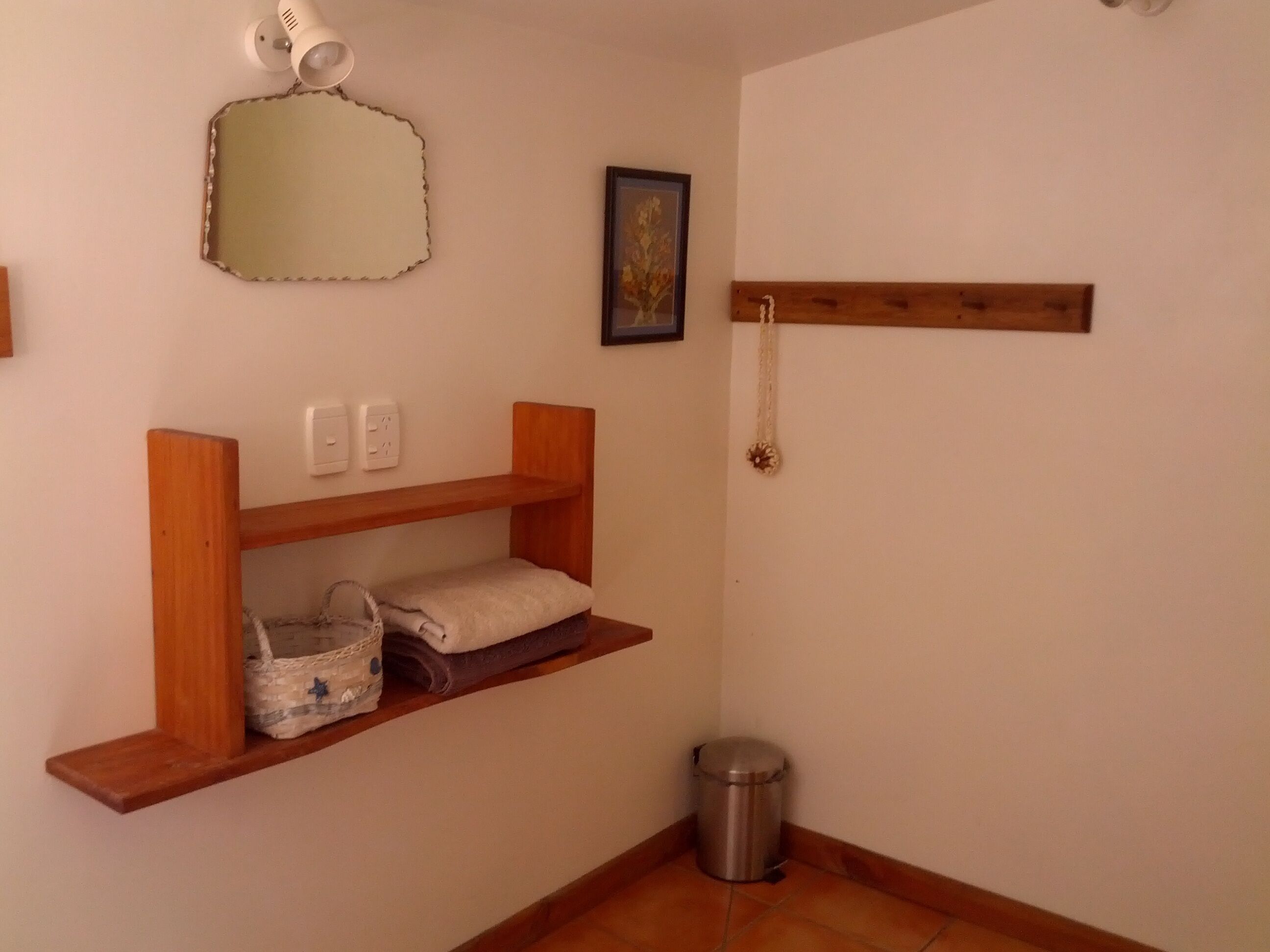 Une salle de bain avec des étagères en bois, un miroir, un tableau encadré, une serviette, une poubelle et un luminaire.