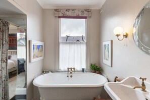 A bathroom with a freestanding bathtub, a sink, and a mirror. There are framed pictures on the wall, a window with curtains, and a door leading to another room. - Lovely 4 Bedroom House near Portobello (London)
