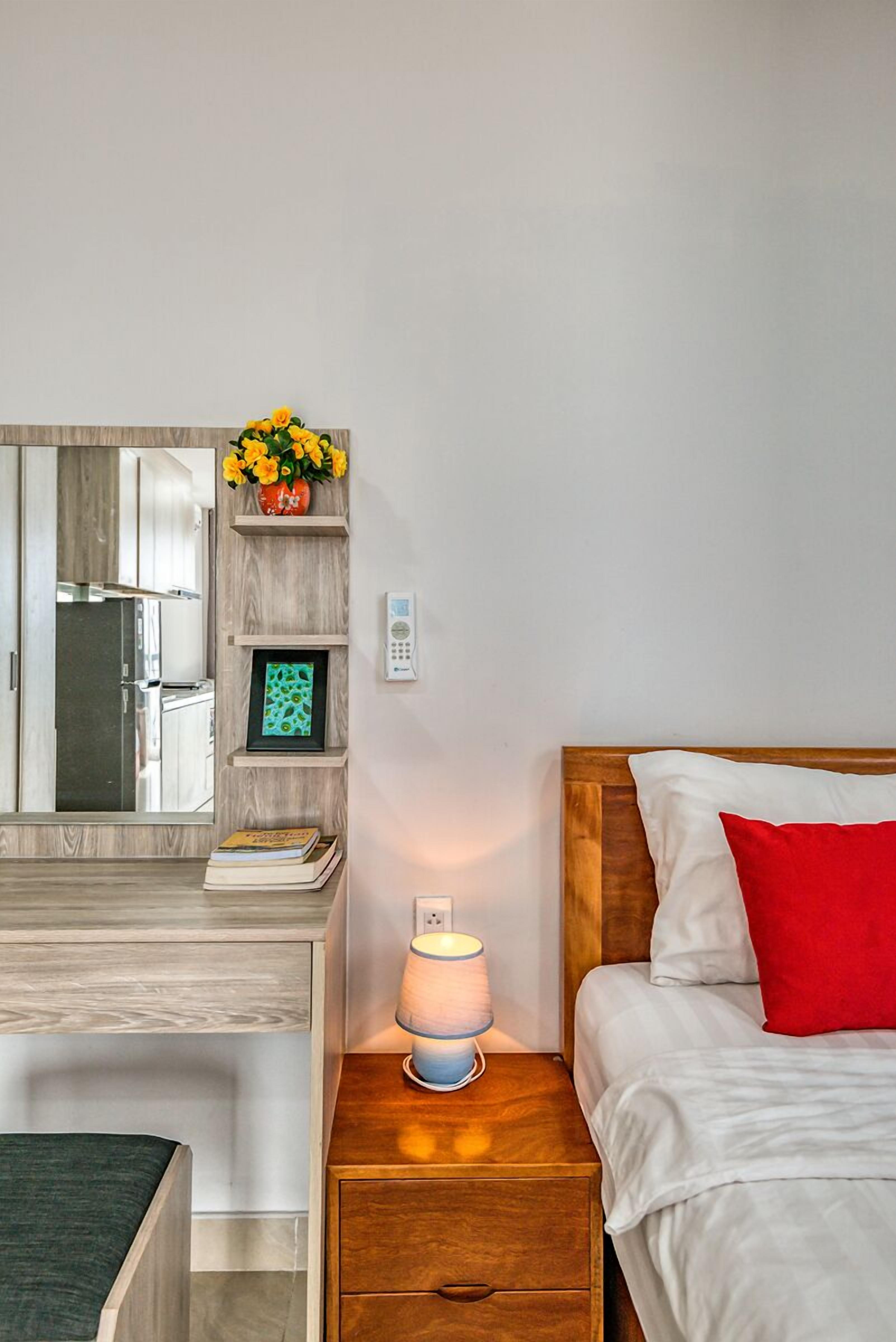 Une chambre à coucher avec une tête de lit en bois, une table de chevet avec une lampe, un oreiller rouge, une commode, un miroir, un tableau encadré et une petite étagère avec des fleurs.