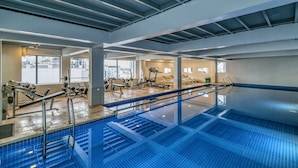 An indoor swimming pool with a clear view of the gym area, exercise equipment, and windows overlooking the city.