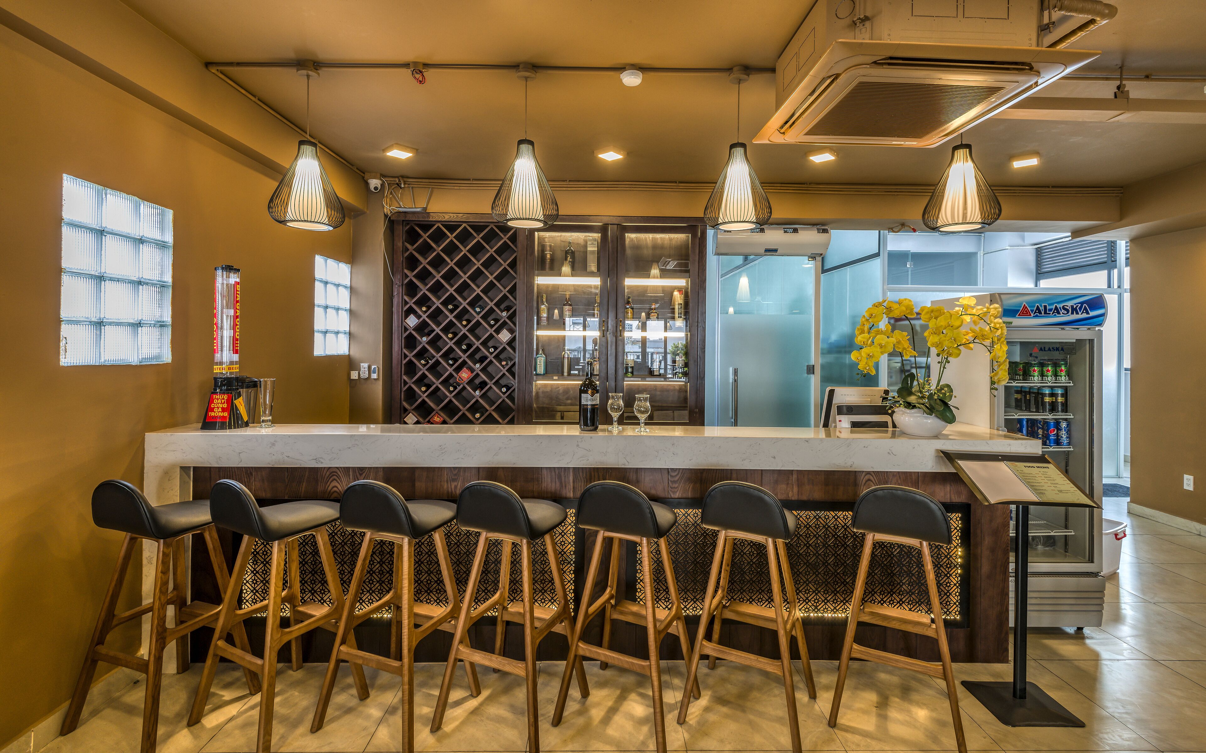 A bar area with a central island, bar stools, hanging pendant lights, a wine cooler, and a refrigerator stocked with beverages.