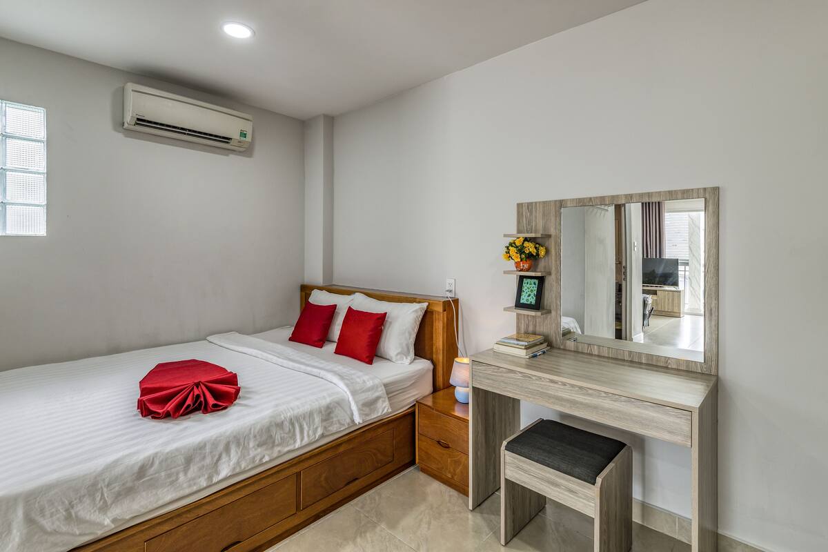 A neatly made bed with white linens and red pillows, a wooden nightstand with a lamp, a mirror, and an air conditioning unit on the wall.