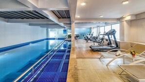 A swimming pool with blue tiles, a gym area with treadmills and weights, and a wooden floor.