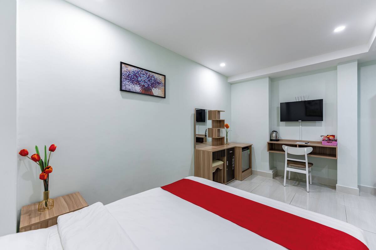 A modern hotel room with a bed, a desk, a TV, and a vase of flowers.