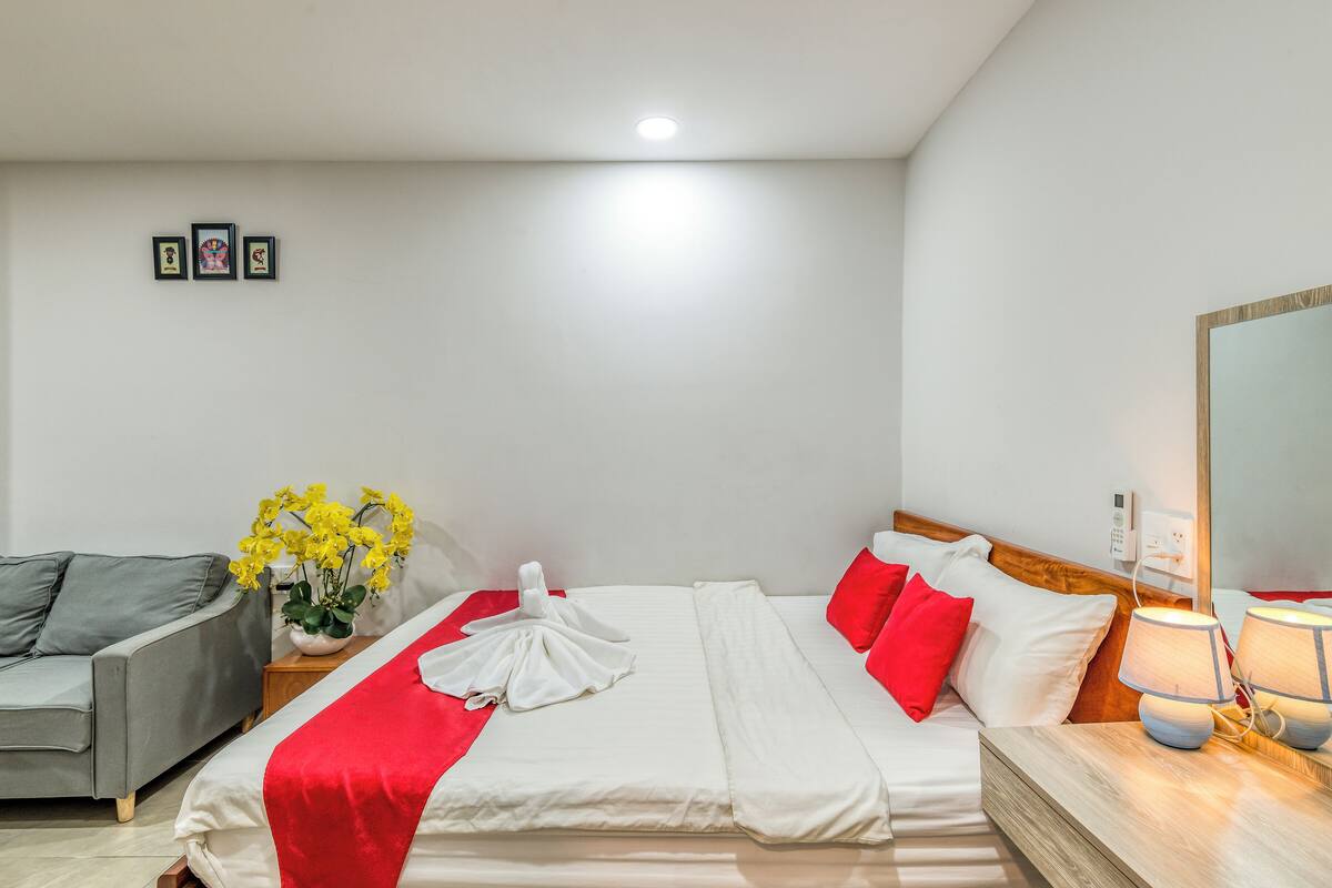A hotel room with a bed, a grey sofa, a vase with yellow flowers, and a bedside table with a lamp.