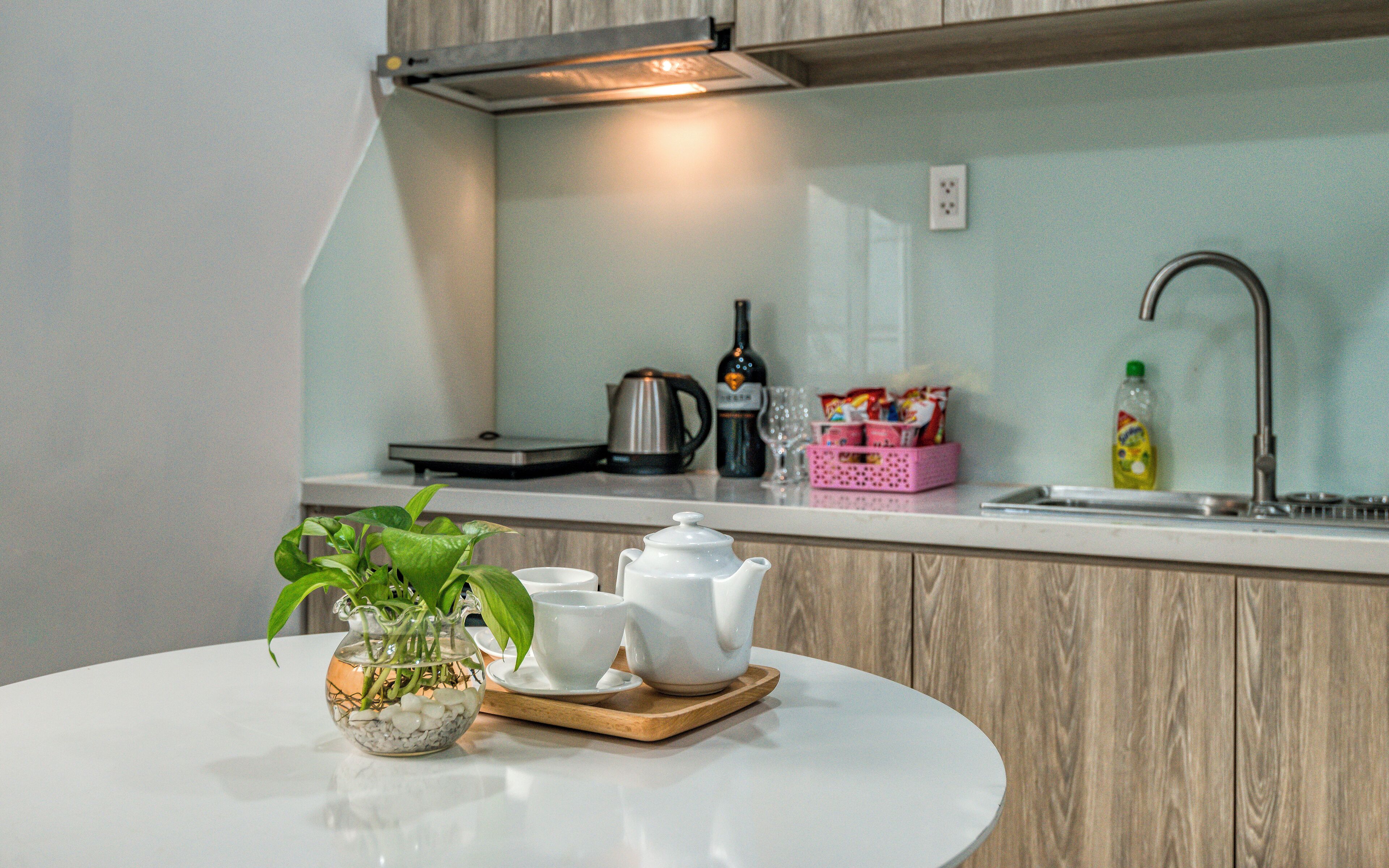 Une cuisine avec un plan de travail blanc, un évier, une bouilloire, une bouteille de vin et une plante dans un vase en verre.