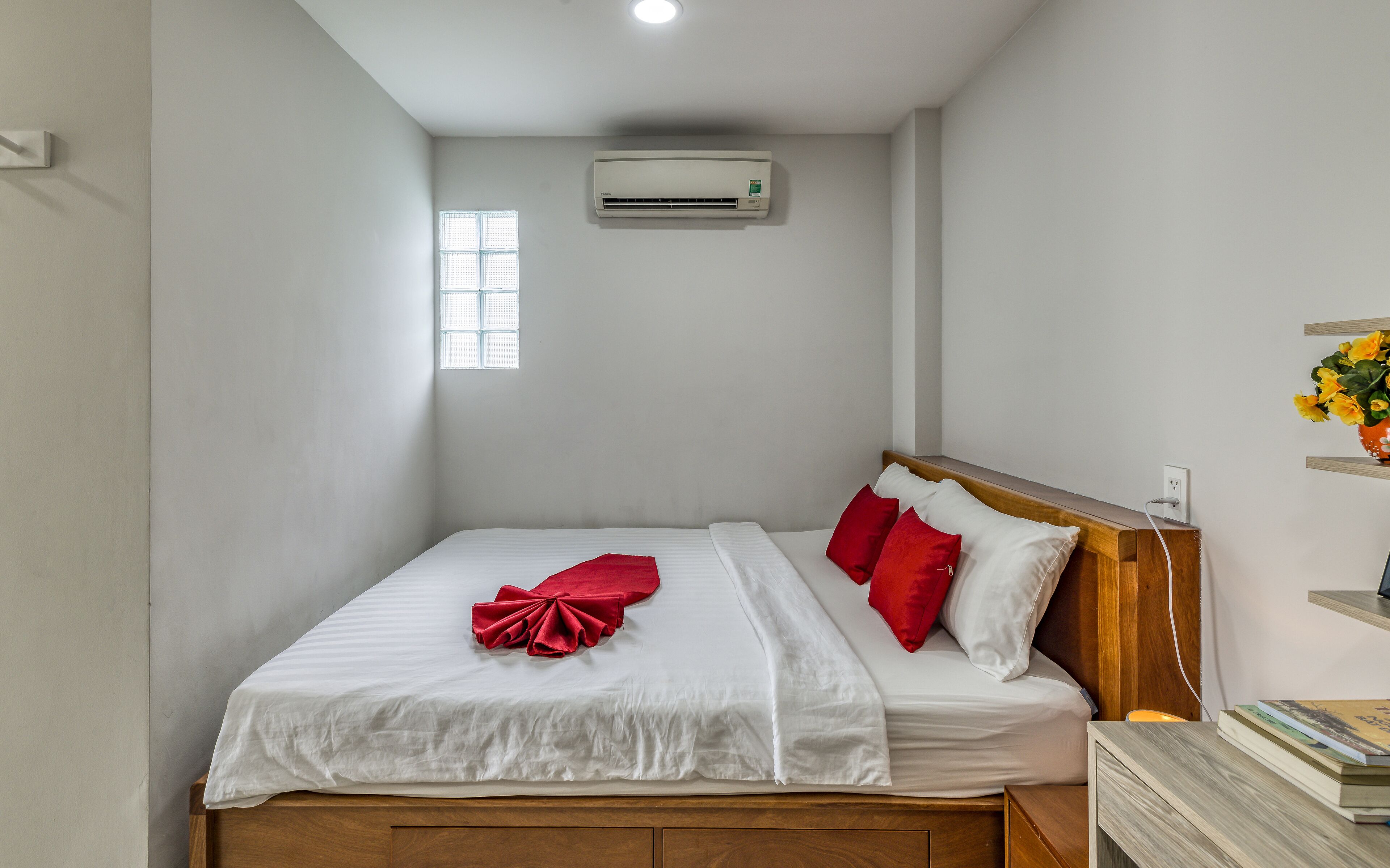 A neatly made bed with white and red bedding, a wooden headboard, and a small window with blinds.