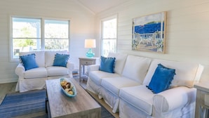 Una habitación luminosa con paredes blancas, dos sofás blancos, cada uno con cojines azules, una mesa de centro de madera y un cuadro con una escena costera.