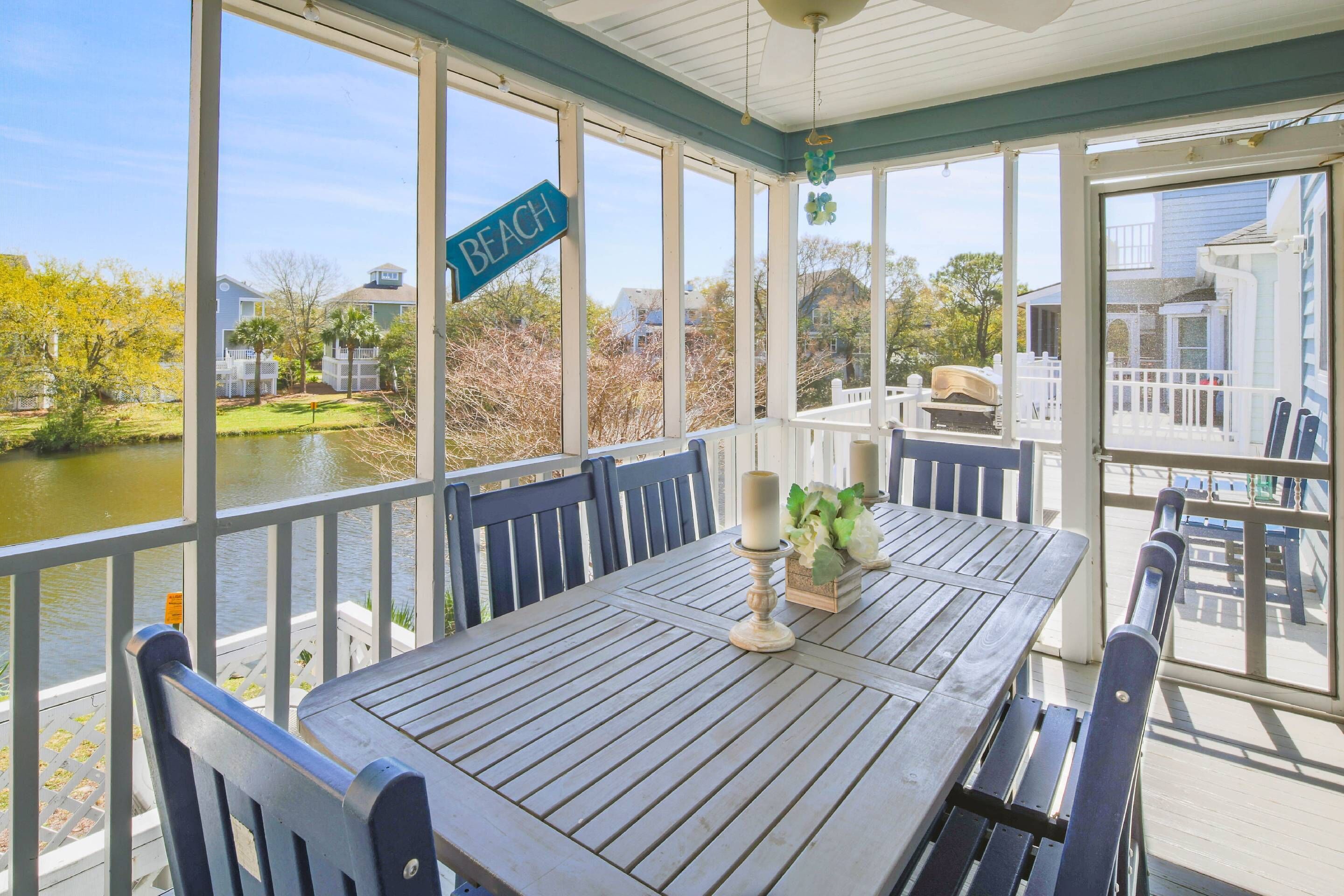 A covered outdoor dining area with a table and chairs, a sign indicating 'BEACH', and a view of a lake and buildings.