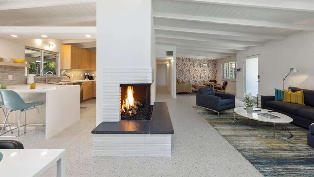 Um interior moderno com lareira central, cozinha com painel de pedra e sala de estar com sofá e mesa de centro.