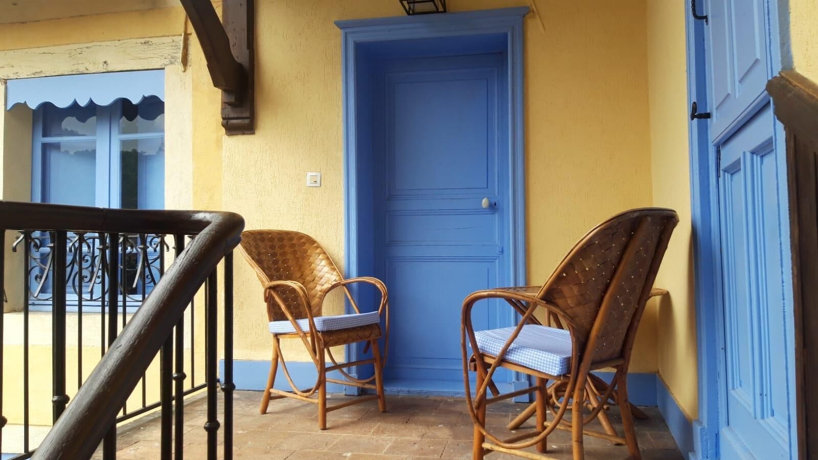 Un balcón con dos sillas de mimbre, una puerta azul y una pared amarilla.