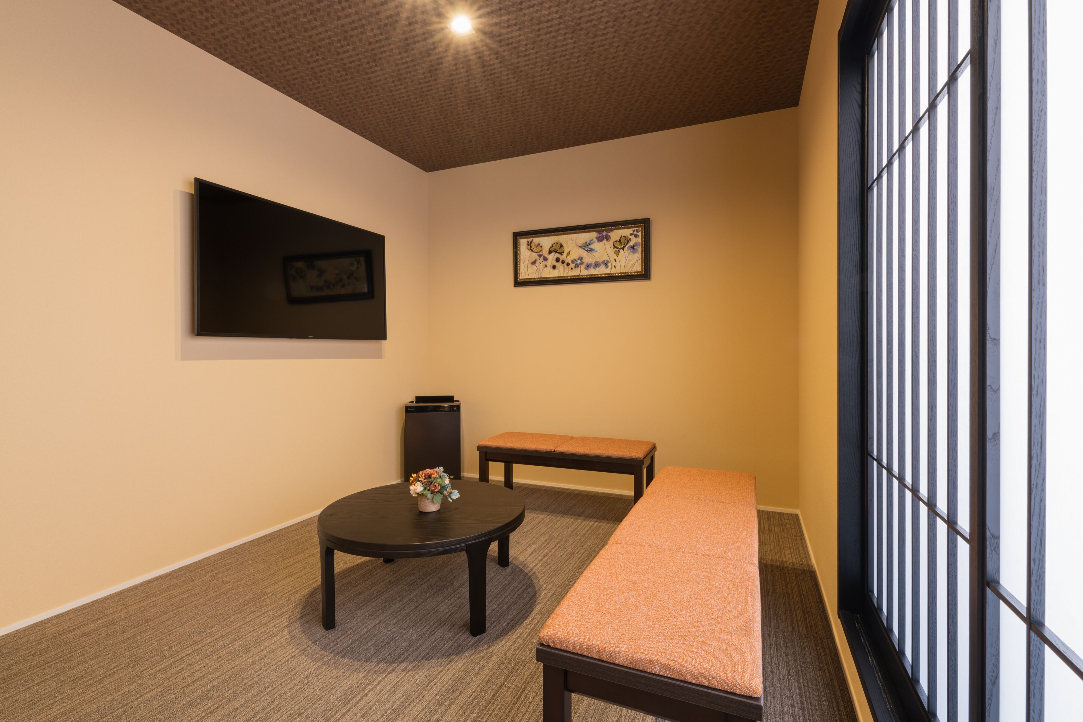 A room with a bed, a small table with a vase, a bench, a TV, and a window with vertical blinds.