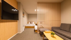 Une chambre d’hôtel moderne équipée d’un téléviseur à écran plat fixé au mur, d’un lit avec une tête de lit en bois, d’un canapé gris, d’une table basse en bois agrémentée d’un vase à fleurs et d’une cloison coulissante en bois.