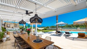 Una mesa de comedor de madera con sillas de mimbre, una lámpara colgante y vistas a una piscina y follaje.
