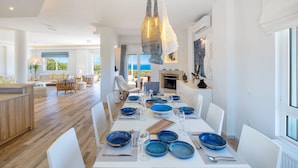 Un comedor amplio con una mesa larga preparada para una comida, una cocina al fondo y vistas al exterior.