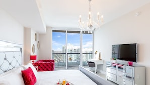 A modern hotel room with a large bed, a TV, a balcony with a city view, and a desk with a chair.