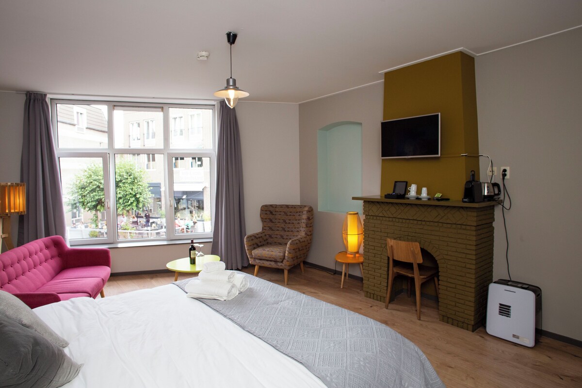 A hotel room with a large bed, a pink sofa, a small round table with a lamp, a brick fireplace, and a television mounted on the wall.