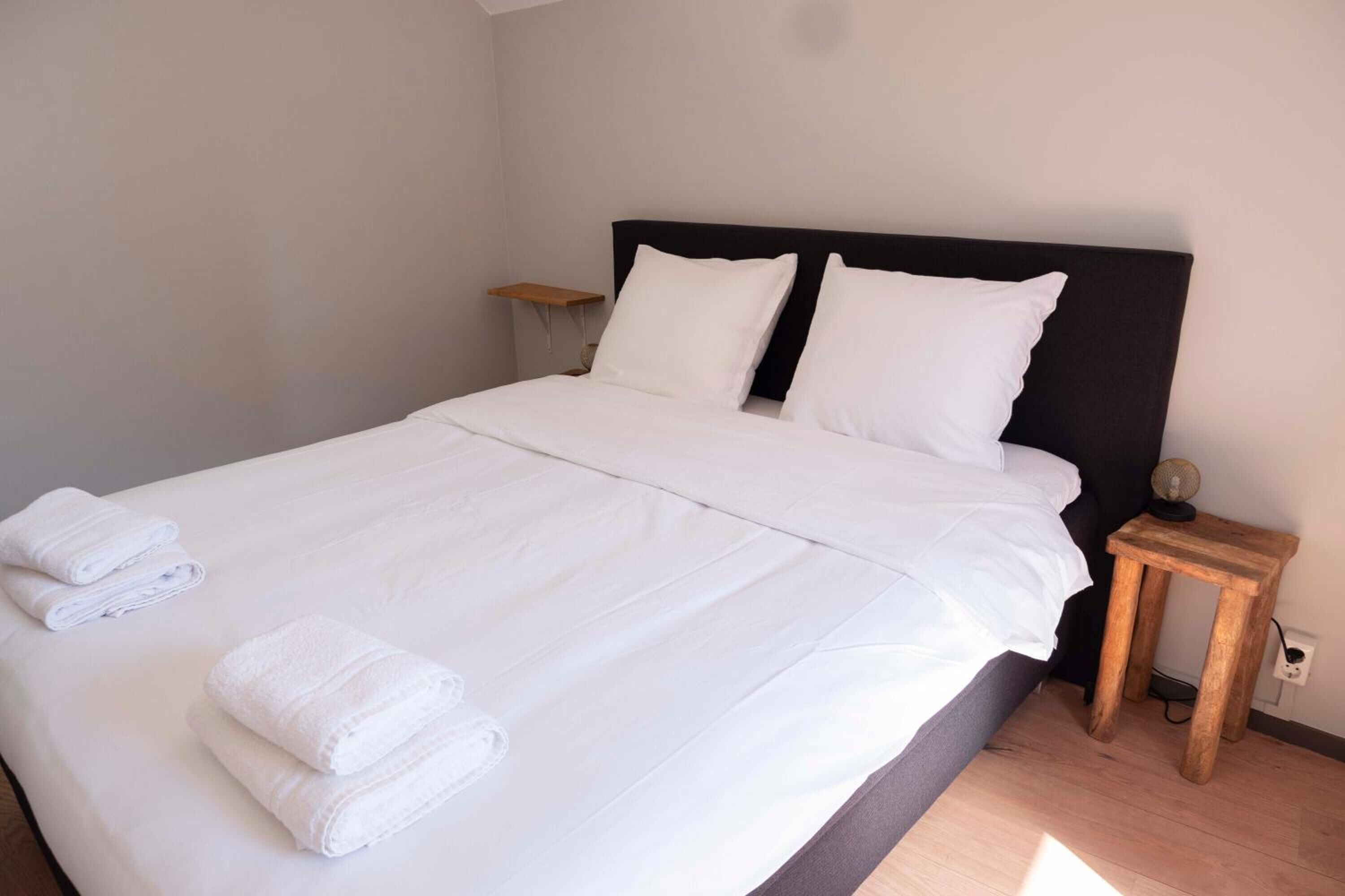 Un lit bien fait, avec du linge de lit et des serviettes blanches, une table de chevet en bois avec une lampe, et une petite table de nuit dotée d’une prise électrique.