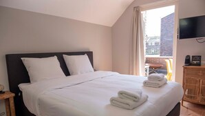 A neatly made bed with white linens and towels, a wooden nightstand, a television on the wall, and a balcony with a view of buildings. - Holt Nijmegen (Nijmegen)