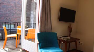 Una habitación con una silla azul, una silla naranja, una pequeña mesa con libros y un televisor en la pared.