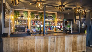 Uma área de bar com balcão de madeira, prateleiras com garrafas de bebidas e luminárias pendentes.
