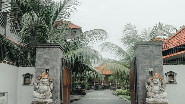 A pathway leading to a building with a tiled roof, flanked by palm trees and statues. - Hotel Diana 2 (Denpasar)