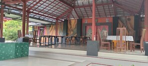 A covered outdoor area with a tiled floor, wooden chairs, a table with a white tablecloth, and a green tablecloth with a leaf pattern. - Penginapan Pendopo Kasih (Semin)