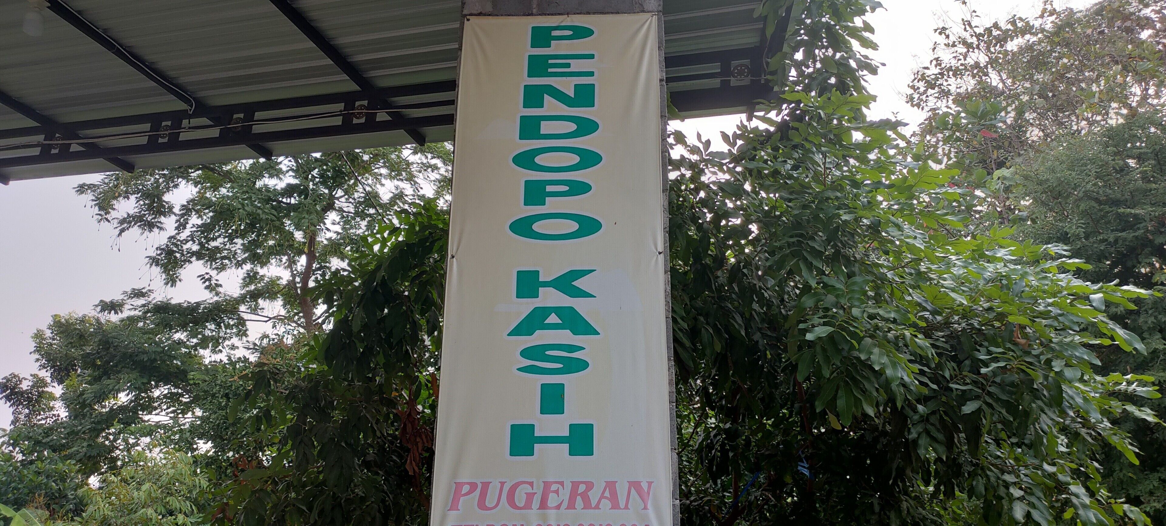 A banner with the text "PENDOPO KASH PUGERAN" hanging under a roofed structure surrounded by trees.