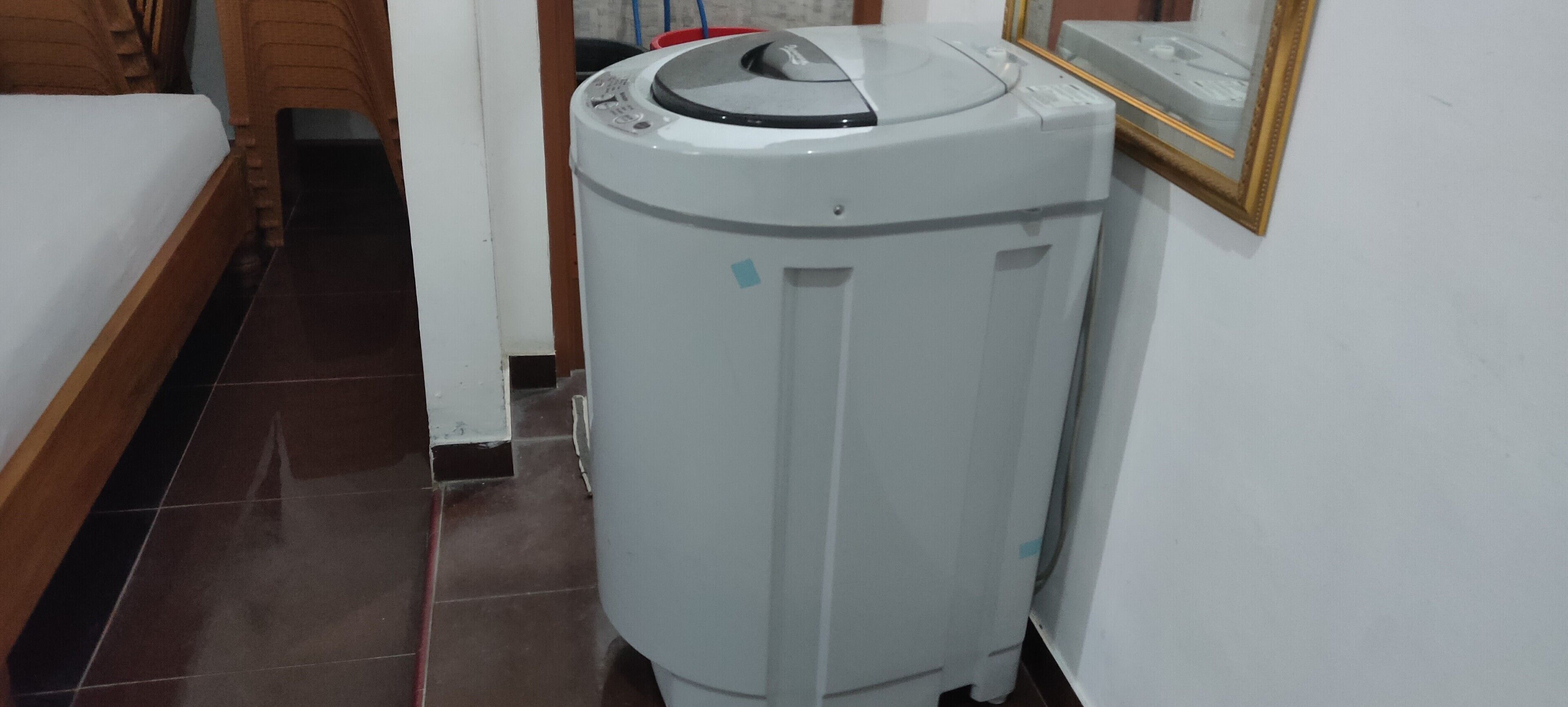 A compact, white, wall-mounted washing machine in a narrow hallway with a wooden bed and a tiled floor.