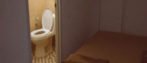 A small, dimly lit room with a single bed and a toilet visible through a doorway.