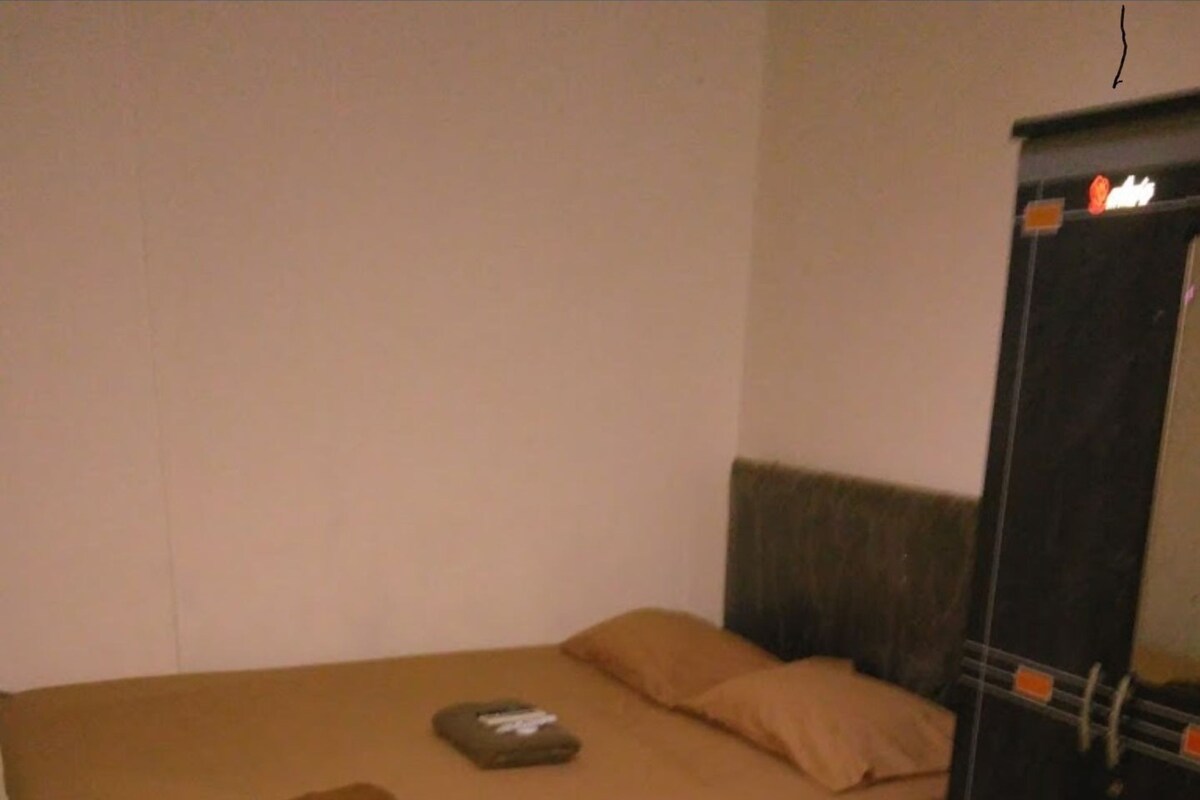 Una cama individual con colchón y almohadas marrones, un objeto pequeño doblado sobre la cama, y un armario negro con detalles anaranjados.