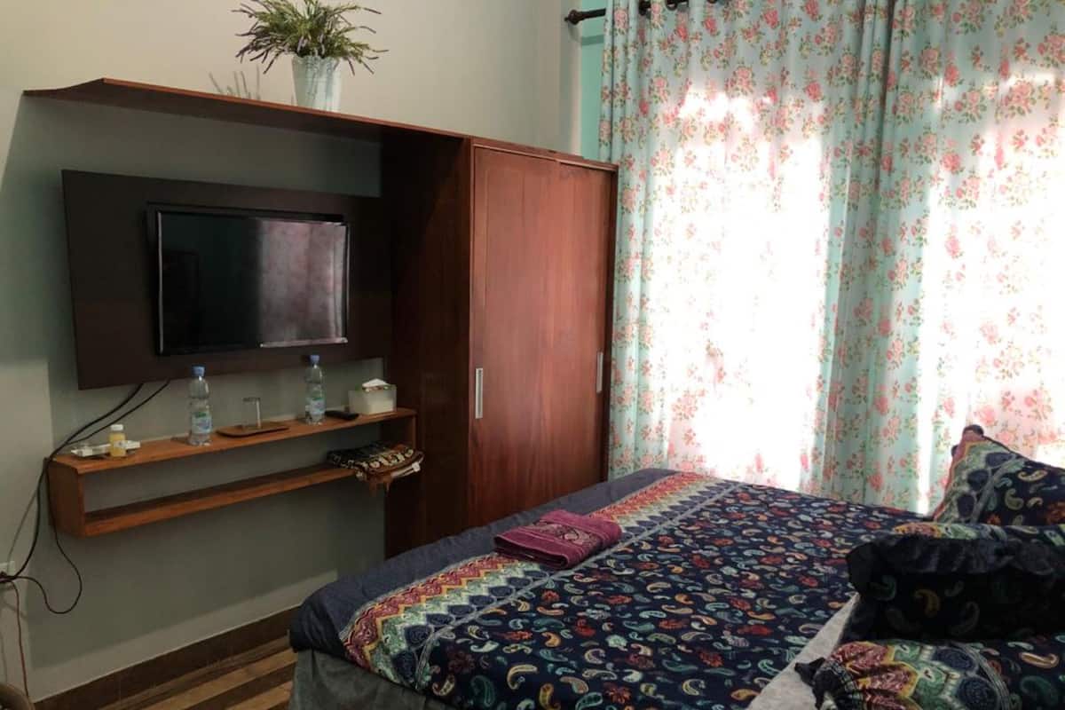Ein Schlafzimmer mit einem Bett, einem an einer Holzhilfe montierten Fernseher, einem Kleiderschrank und einem Fenster mit blumigen Vorhängen.