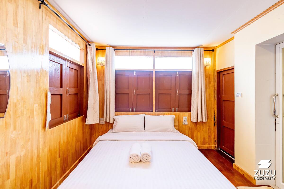 A hotel room with wooden walls, a large bed with white linens, two windows with curtains, and a door on the right.