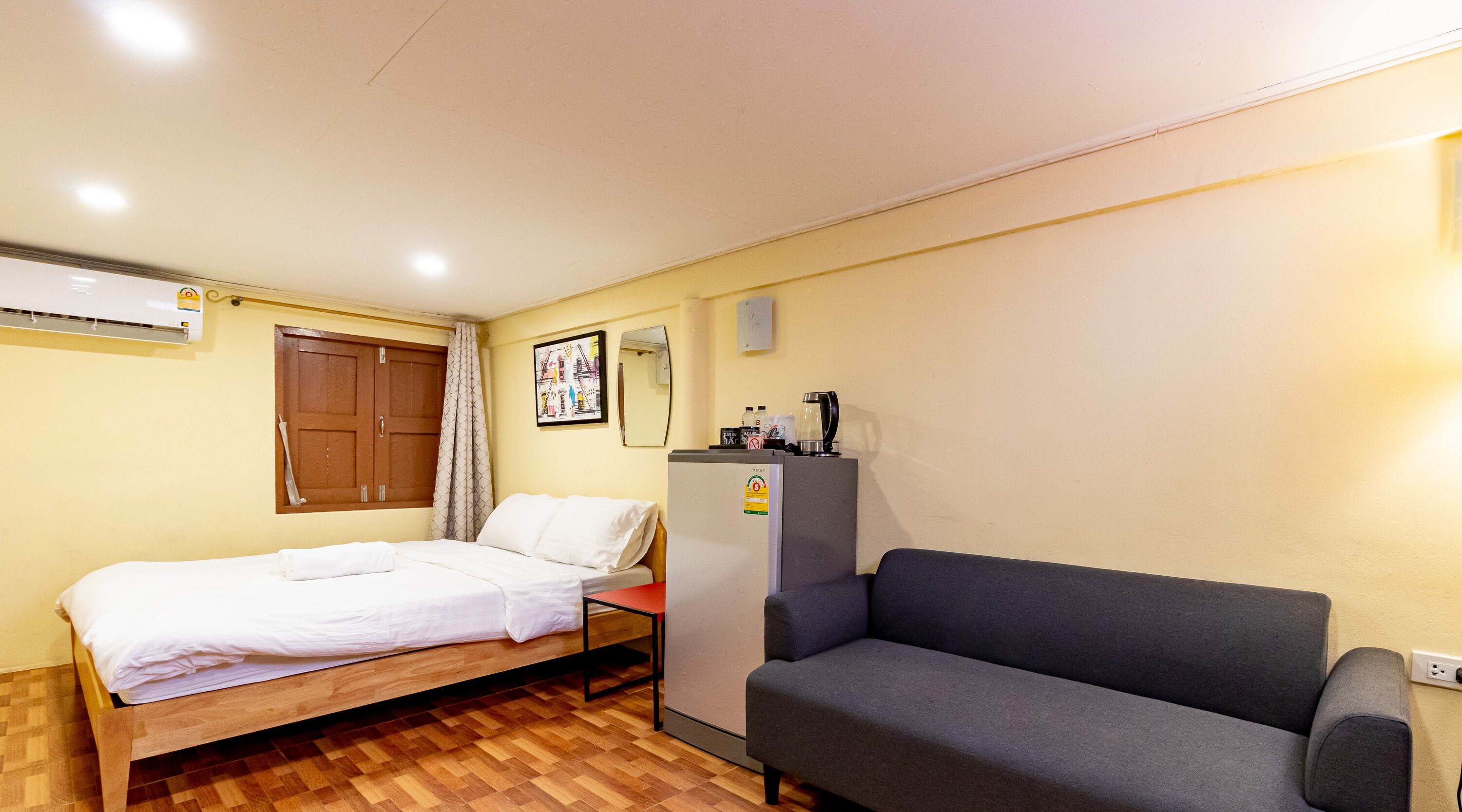 A hotel room with a wooden bed, a grey sofa, a refrigerator, and a wall-mounted air conditioner.