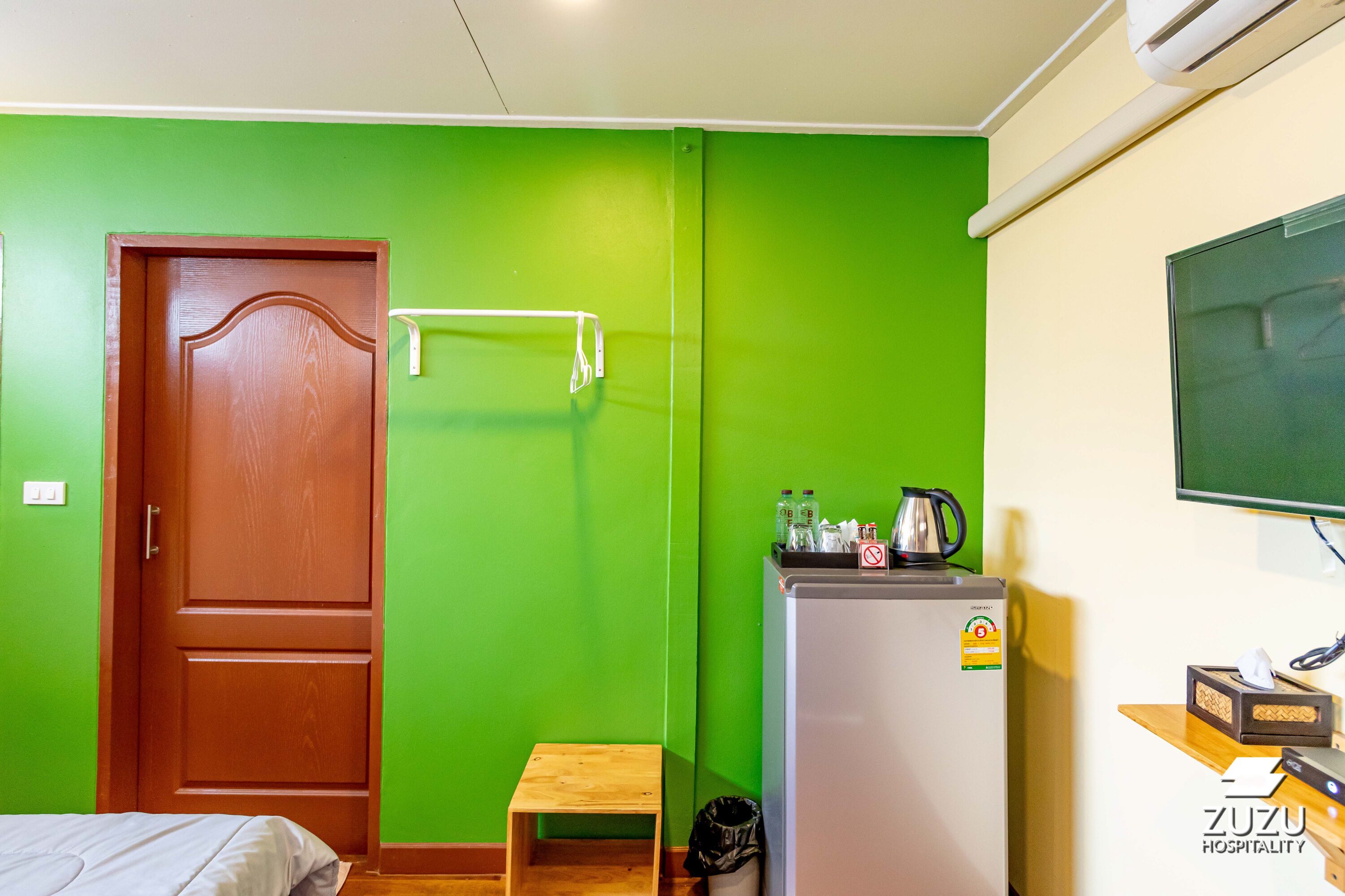 Habitación de hotel con pared verde, puerta de madera, taburete pequeño de madera, refrigerador y televisor montado en la pared.