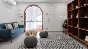 A modern living room with a blue sofa, two round ottomans, a patterned rug, a wooden bookshelf, and a large arched window.