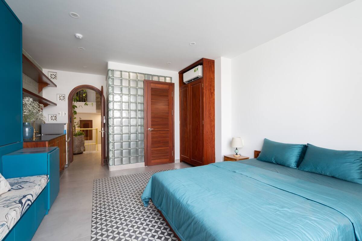 Een moderne slaapkamer met een bed, een blauwe bank, een houten garderobe en een keuken in de achtergrond.