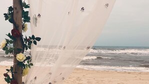 Ein Hochzeitbogen am Strand mit weißem Stoff und Blumendekorationen.