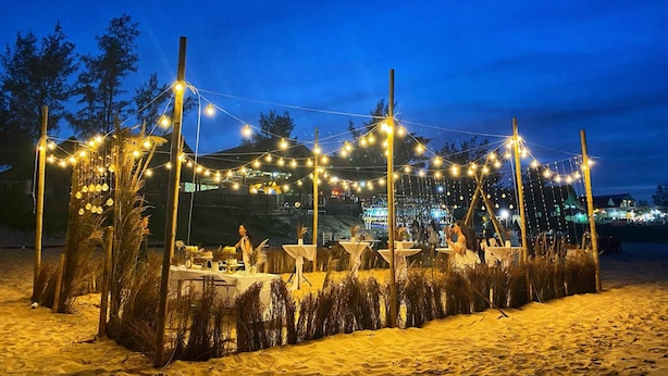 Eine Open-Air-Veranstaltung mit Tischen auf sandigem Untergrund, dekoriert mit Lichterketten und umgeben von Bambusstangen.
