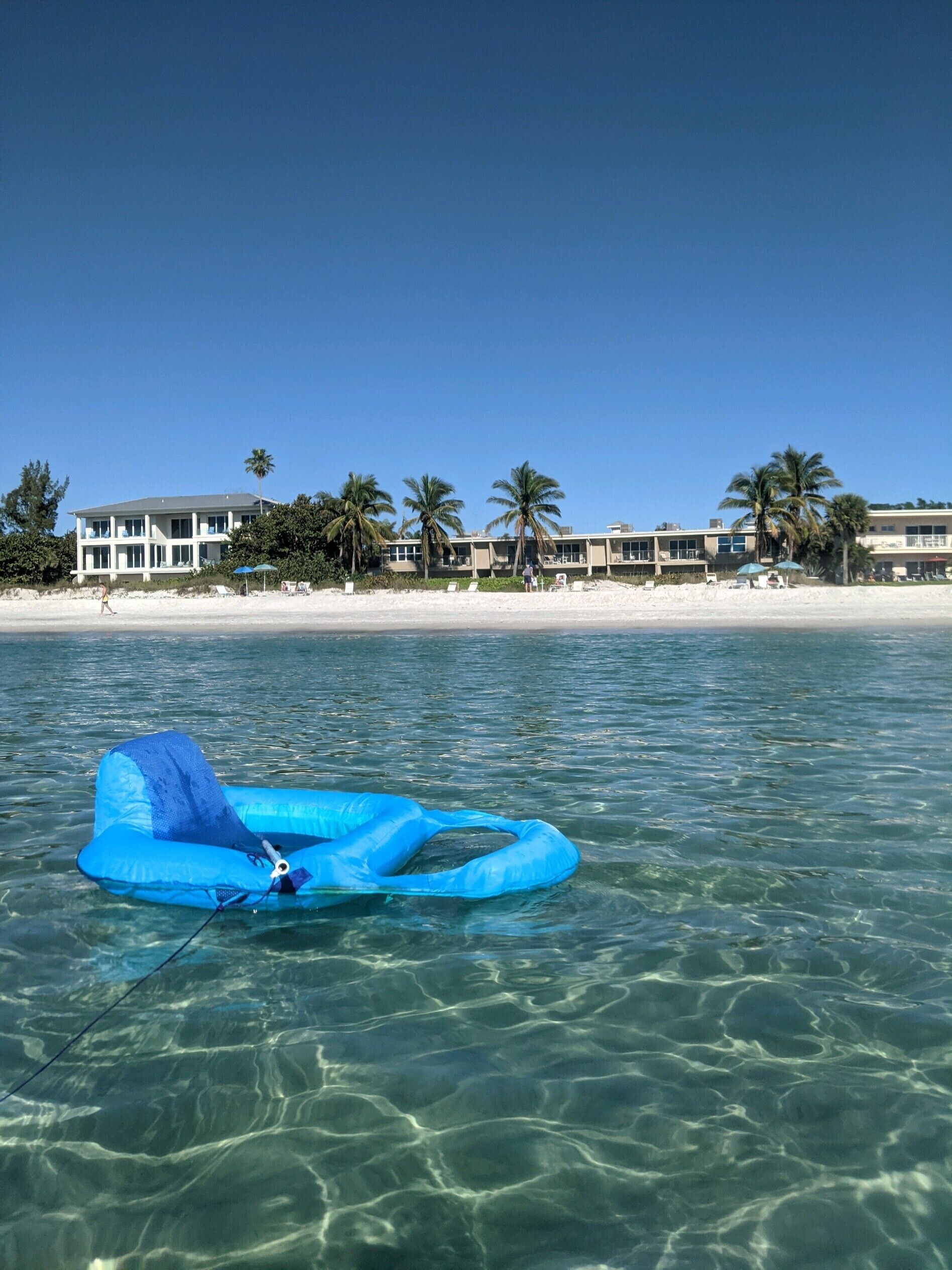 Une plage dégagée, avec un radeau gonflable bleu flottant sur l’eau, des palmiers et, au fond, un bâtiment doté de nombreuses fenêtres.