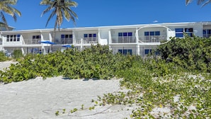 Un hotel bianco a più piani con balconi, situato vicino a una spiaggia con palme e vegetazione lussureggiante.